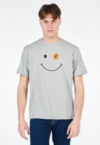 T-shirt in cotone grigio con una grande grafica di un sorriso nero e una patch circolare arancione con la silhouette di un'anatra. Maniche corte, colletto rotondo.
