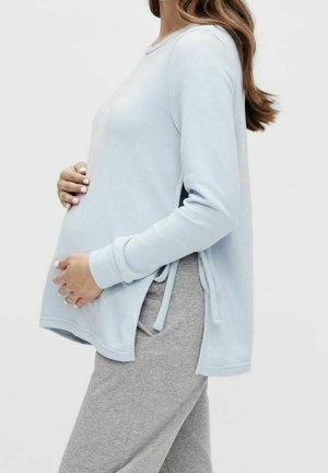Gravid kvinde iført lyseblå langærmet top og grå bukser, der blidt holder sin mave og står sidelæns mod en ensfarvet baggrund.