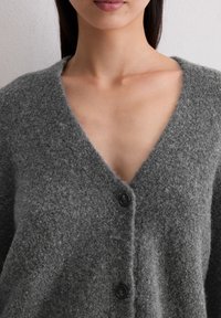 Grijze wollen cardigan met een V-hals, zachte textuur en drie zwarte knopen aan de voorkant. De stof lijkt dik en comfortabel.