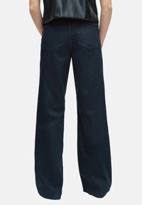 Osoba nosząca ciemnoniebieskie dżinsy z wysokim stanem i szerokimi nogawkami, z tylnymi kieszeniami, stoi na jednolitym jasnym tle, plecami zwrócona w stronę widza.