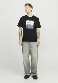 Černé bavlněné tričko s grafikou „JACK & JONES“ v bílé a šedé barvě, spárované se svetrovými modrými volnými džínami a černými botami.