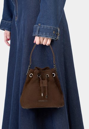 Sac seau en suède marron avec fermeture à cordon, ferrures argentées et poignée courte, porté avec une robe en jean. Nom de la marque visible à l'avant.