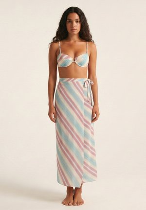 Mujer de pie descalza con un top de bikini de rayas en tonos pastel y una falda larga envolvente de talle alto a juego, sobre un fondo liso.