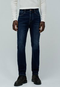 Jeans de mezclilla azul oscuro de corte ajustado, con costuras en contraste, cinco bolsillos y cierre de botón metálico. Combinados con botas de cuero negras.
