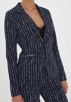 Vrouw draagt een marineblauw pak met witte verticale gestippelde strepen, zwart kanten topje zichtbaar onder het één-knops colbert.