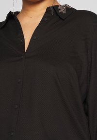 Camicia nera con un motivo testurizzato, apertura frontale con bottoni e colletto ricamato dettagliato. Presenta una vestibilità ampia e un tessuto morbido.
