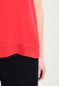 Top smanicato a strati rossi indossato con pantaloni neri, che mostra il lato destro del torso e del braccio di una persona su uno sfondo chiaro.