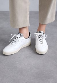 Zapatillas de cuero blancas con diseño perforado, acentos en negro y estampado de serpiente en el cuello, y una suela texturizada de color crema.