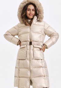 Beige puffer coat with a hood featuring faux fur trim, quilted design, front zipper, belted waist, and large side pockets.