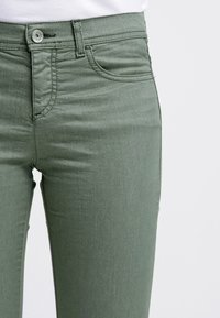 Olivevihreät farkut, jotka on valmistettu sileästä kankaasta ja joissa on klassinen viisi-taskumalli, vyölenkit sekä edessä nappi- ja vetoketjukiinnitys.