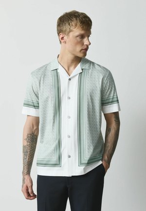 Tânăr cu păr blond scurt poartă o cămașă albă și verde cu mâneci scurte cu nasturi și pantaloni negri, privindu-se spre dreapta.