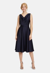 Robe satin bleu marine avec un décolleté drapé, sans manches, et une taille cintrée menant à une jupe évasée, associée à des sandales argentées.