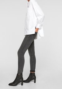 Weißes Hemd mit langen Ärmeln, graue enge Jeans und schwarze Ankle-Boots mit Blockabsatz und seitlichem Reißverschluss.