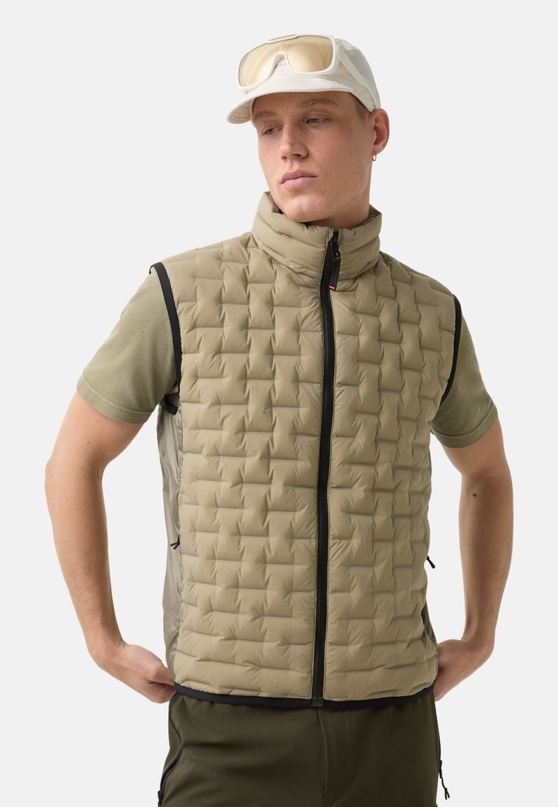 Beżowa ocieplana kamizelka w diamentowy wzór, z wysokim kołnierzem, suwakiem z przodu i czarnymi akcentami. Noszona na oliwkowej koszuli z krótkim rękawem, slim fit.