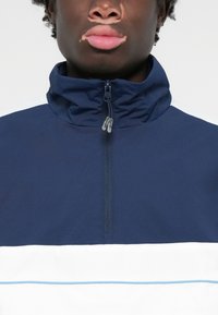 Marineblå og hvid zip-jakke med høj krave; har et let stof og en horisontal hvid stribe med en lyseblå akcent.