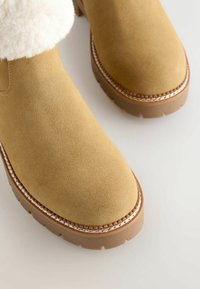 Bruna mockaskor i ankellängd med en tjock vit pälskrage, med kontrasterande sömmar och en texturerad gummisula med djupa mönster.