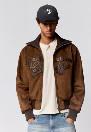 Junger Mann trägt eine braune Jacke mit Reißverschluss und Text, ein weißes strukturiertes Henley-Shirt, blaue Jeans und eine schwarze Kappe mit der Aufschrift "Ny".
