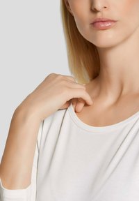 Weißes Baumwolloberteil mit rundem Ausschnitt, das eine glatte Stoffstruktur aufweist. Die Hand des Models ruht sanft auf der Schulternaht. Sanfte, natürliche Beleuchtung.