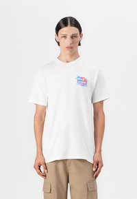 Biała bawełniana koszulka z krótkim rękawem z kolorowym nadrukiem na piersi. Modelka nosi beżowe szorty typu cargo.