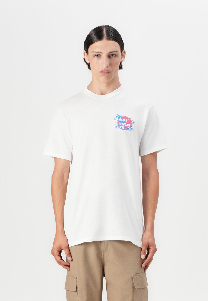 White cotton t-shirt with short sleeves, featuring a colorful graphic print on the left chest. The model is wearing beige cargo pants.