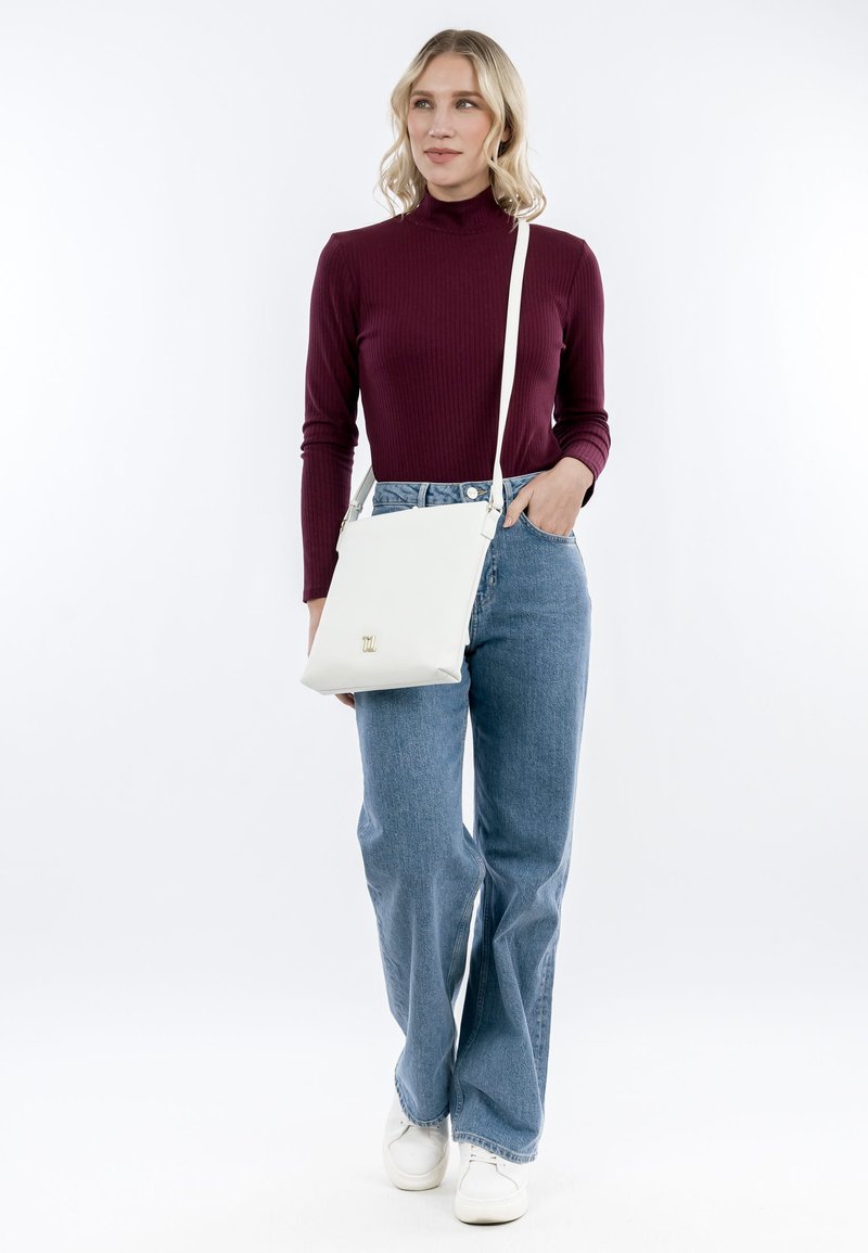 Frau in burgunderrotem Langarmshirt und blauen Jeans, mit weißer Umhängetasche und weißen Sneakers, steht vor hellem, einfarbigem Hintergrund.