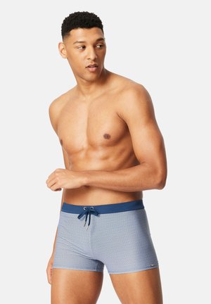 Mann, der blaue Badehose mit Muster, navy-blauem Bund und Kordelzug trägt, steht vor schlichtem, hellem Hintergrund und schaut seitlich weg.