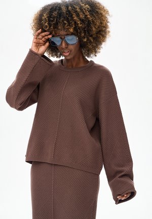 Bruine, textuurvolle oversized trui met een geribde halslijn en losse mouwen, gecombineerd met een bijpassende rok. Model draagt blauwe zonnebril.