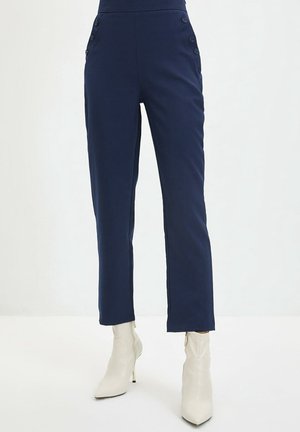 Marineblauwe broek met hoge taille en knoopdetails aan de zijkanten, gecombineerd met witte enkellaarsjes met puntige neus en smalle hakken.