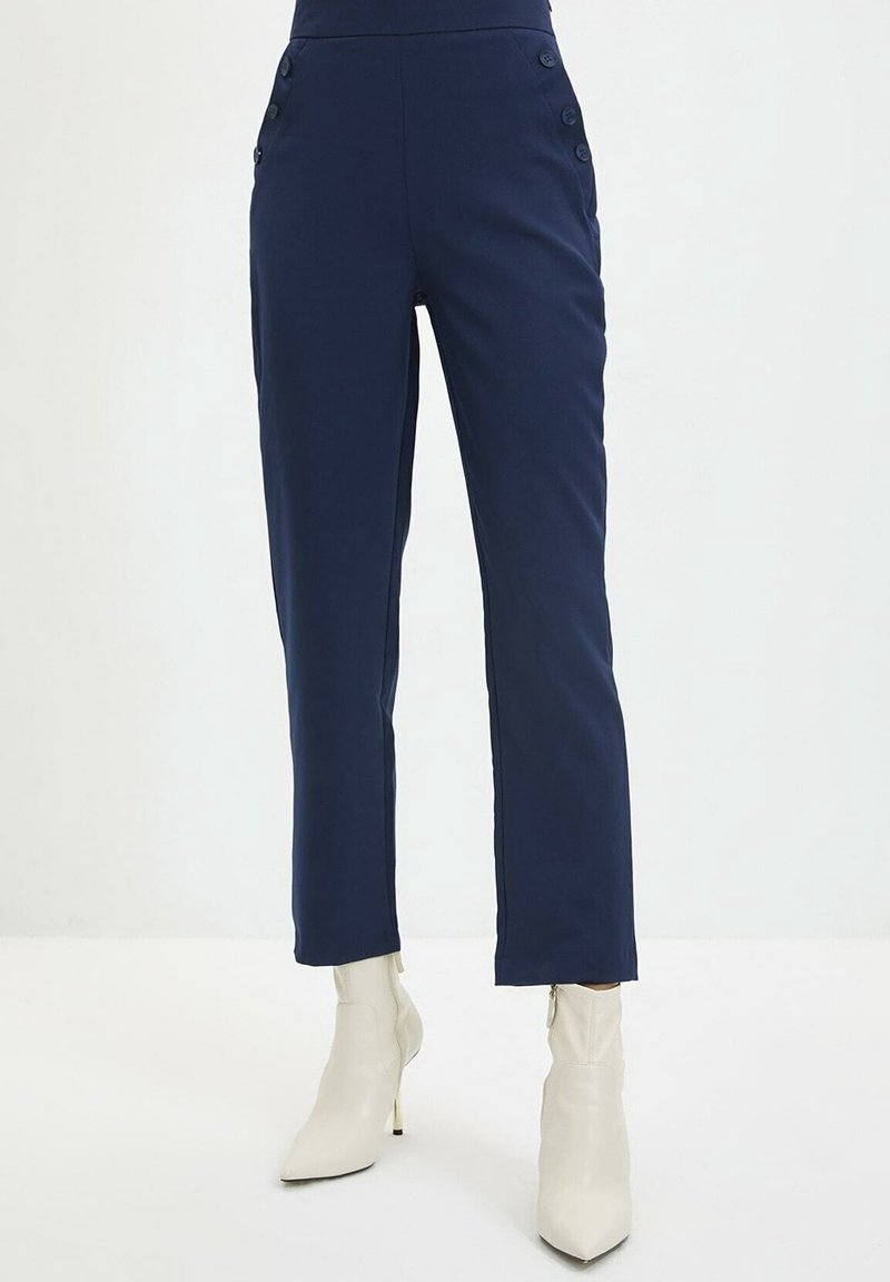 Pantalon taille haute bleu marine avec détails de boutons sur les côtés, associé à des bottines blanches à bout pointu et talons fins.