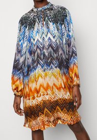 Färgglad klänning med zigzag-mönster, långa ärmar, hög krage och rynkdetaljer. Har nyanser av blått, orange, brunt och vitt.
