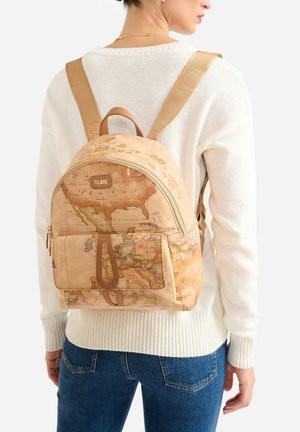 Donna che indossa un maglione bianco e jeans blu con zaino marrone chiaro decorato con una mappa vintage del mondo e una tasca frontale.