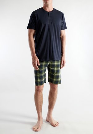 Ein Mann steht barfuß und trägt ein marineblaues Henley-Shirt mit kurzen Ärmeln sowie grün karierte, knielange Shorts vor einem weißen Hintergrund.