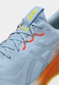 Ανοιχτό μπλε παπούτσι τρεξίματος Asics Gel-Cumulus 22 με κίτρινο λογότυπο στη γλώσσα, πορτοκαλί θηλιές κορδονιών και σόλα με βαθμιαία μετάβαση από πορτοκαλί σε κίτρινο.