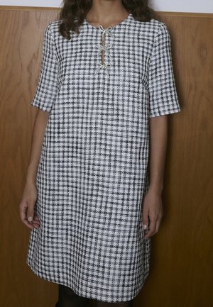 Femme portant une robe à carreaux noir et blanc à manches courtes avec deux nœuds papillon au niveau de l'encolure, debout devant un fond en bois.