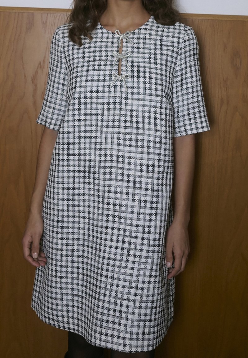 Femme portant une robe à carreaux noir et blanc à manches courtes avec deux nœuds papillon au niveau de l'encolure, debout devant un fond en bois.