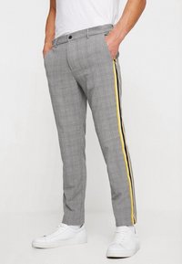 Šedé kostkované kalhoty s úzkým střihem, ozdobené žluto-černými pruhy po stranách, s hladkou texturou látky. Kombinovány s bílými teniskami.