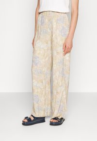 Kvinna som bär beige vida byxor med pastellfärgad blommönster och svarta plattformssandaler mot en vit bakgrund.