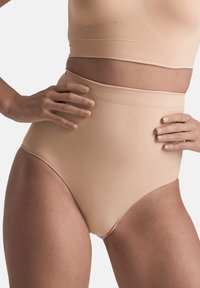 Donna che indossa una guaina modellante beige a vita alta e un reggiseno abbinato, con le mani sui fianchi su uno sfondo neutro.