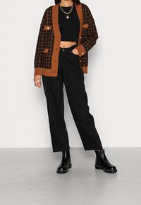 Una donna indossa un top corto nero, un cardigan a scacchi marrone e nero, pantaloni neri a vita alta, stivaletti neri e collane d'oro a più strati.