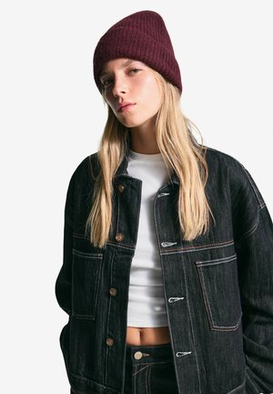 Black denim jacket with large pockets, light stitching, and bronze buttons. White t-shirt underneath and maroon knitted beanie on head.