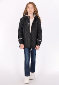 Svart vattentät jacka med grå huva, med tryckknappar, elastiska ärmslut och reflexdetaljer, bärs över jeans och vita sneakers.