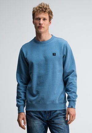 Blue crewneck sweatshirt made of soft fabric, featuring ribbed cuffs and hem, with a small black logo patch on the left chest.