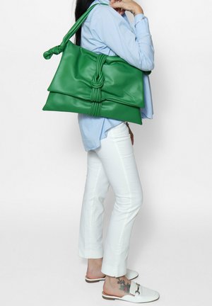 Borsa a tracolla in cuoio verde con manico annodato e due sezioni sovrapposte. Texture liscia, grande dimensione, portata sulla spalla contro una camicia chiara.