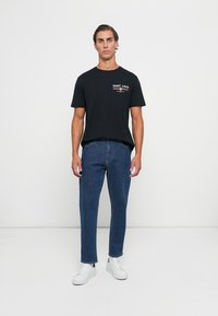 Ung mand iført en sort GANT 1949 t-shirt, blå jeans og hvide sneakers, der står imod en ensfarvet hvid baggrund.