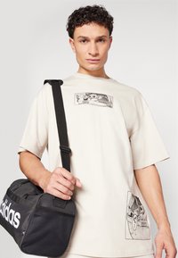 Beige cotton t-shirt featuring two graphic prints, one on the front and one on the side. Black duffel bag with white logo, held by a short strap.