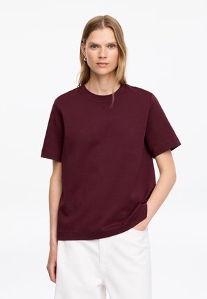 Femme aux cheveux blonds raides porte une chemise rouge foncé à manches courtes et un pantalon blanc, avec une main dans la poche, sur un fond uni.