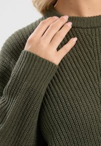 Olivgrüner, gestrickter Pullover mit Rippstruktur. Nahaufnahme einer Hand, die auf der Schulter ruht und das Detail des Kragens und der Ärmel zeigt.