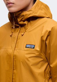 Gros plan d'une personne portant une veste Patagonia jaune moutarde avec une capuche et le logo visible de la marque sur la poitrine.