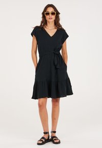 Zwarte jurk tot de knie met een V-hals, korte mouwen, een cintuur in de taille, een geplooide rok en een gerimpelde zoom. G gedragen met zwarte sandalen en een zonnebril.