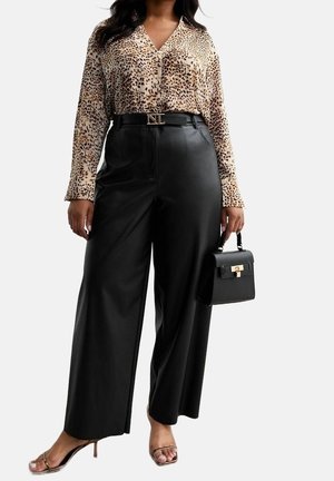 Kvinde iført leopardprint bluse, sorte vidde bukser med høj talje og bælte, holder en sort håndtaske og har på nogle strop-sandaler.